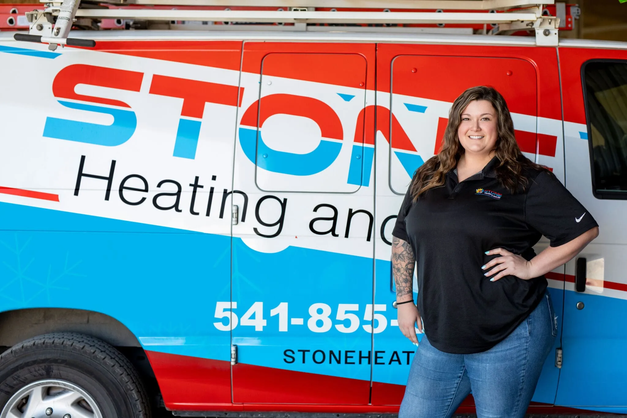 Smiling woman with tattoos stands with hand on hip in front of a red, white, and blue Stone Heating and Air service van.