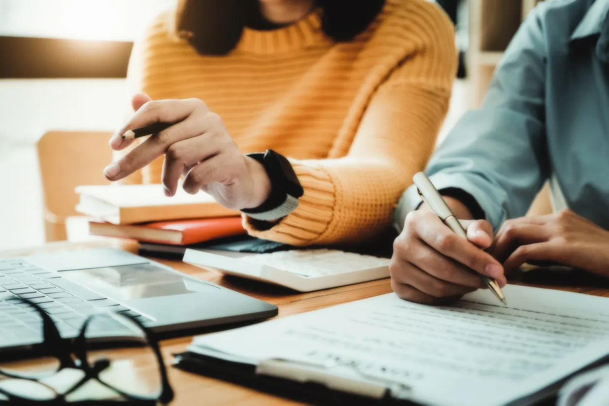 Two people collaborating at a desk with one pointing with a pencil and the other writing on a document.