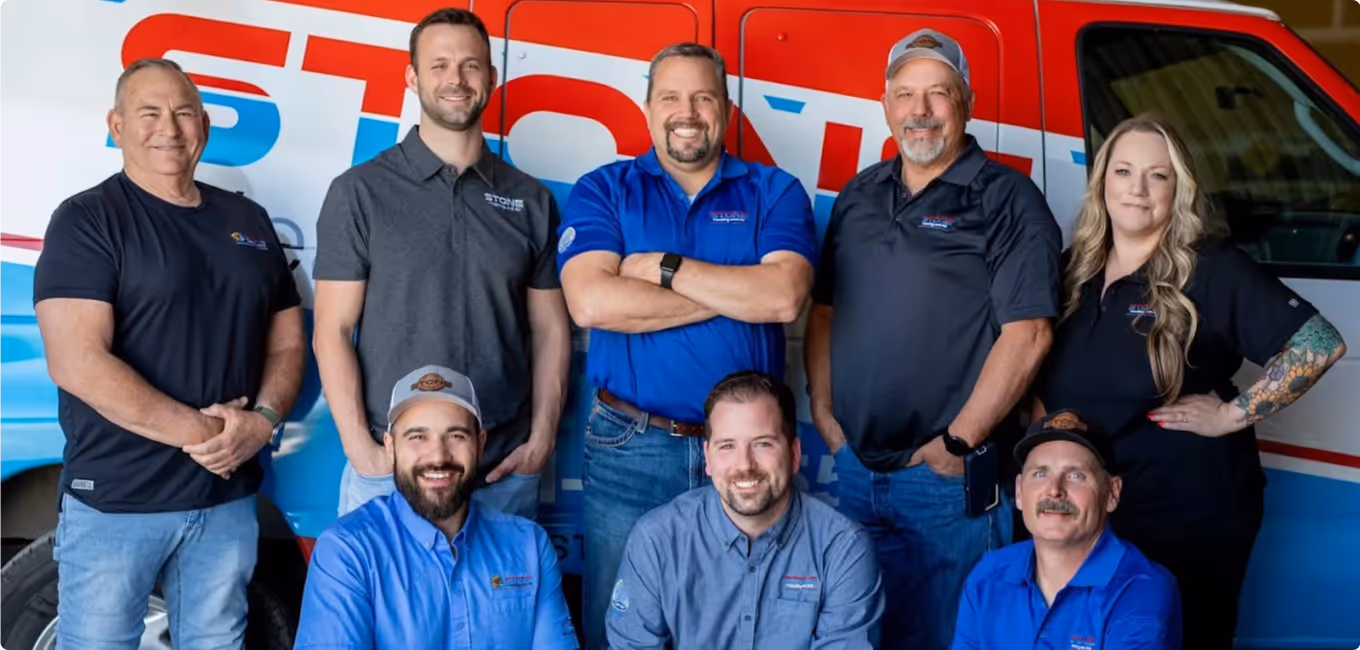 Group of eight diverse professionals in branded work shirts posing and smiling in front of a company vehicle.
