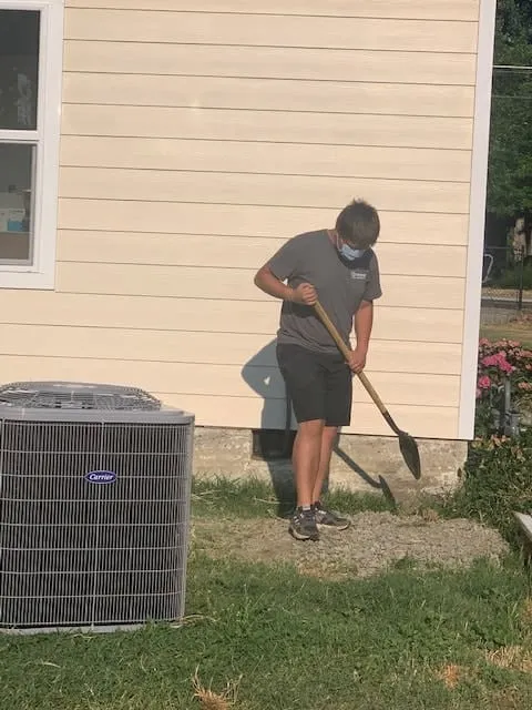 Person wearing a face mask and dark clothes digging with a shovel outside near the corner of a beige house next to an air conditioning unit.