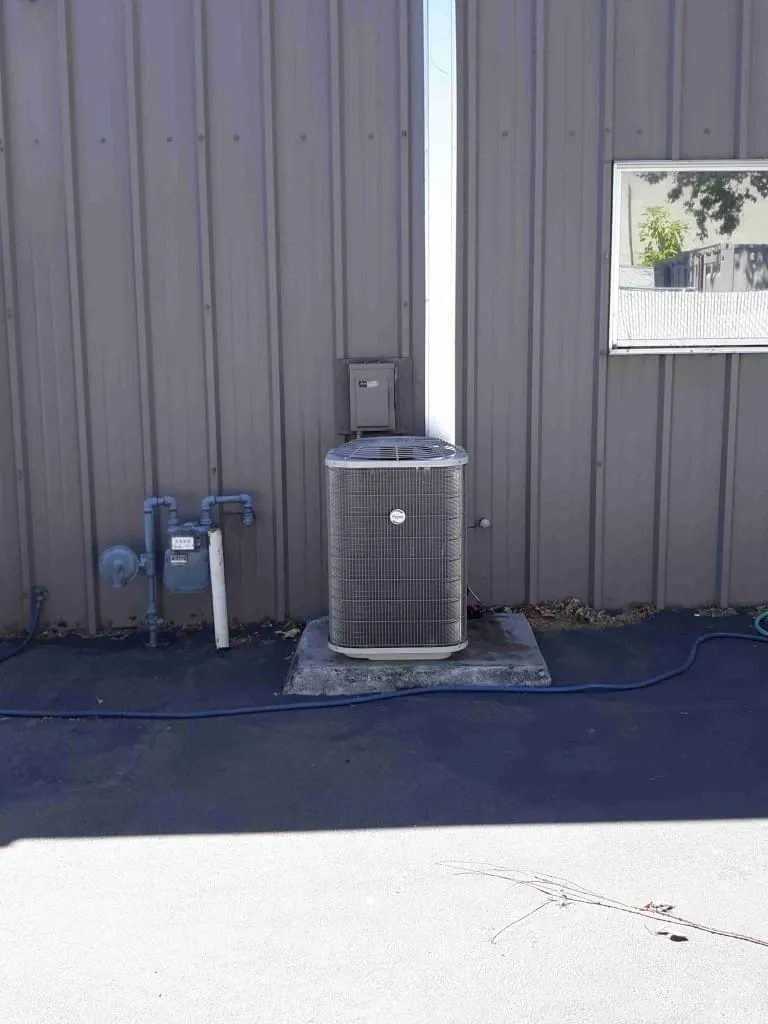 Outdoor air conditioning unit installed on a concrete pad attached to a gray metal building wall with nearby pipes and a window reflecting trees.