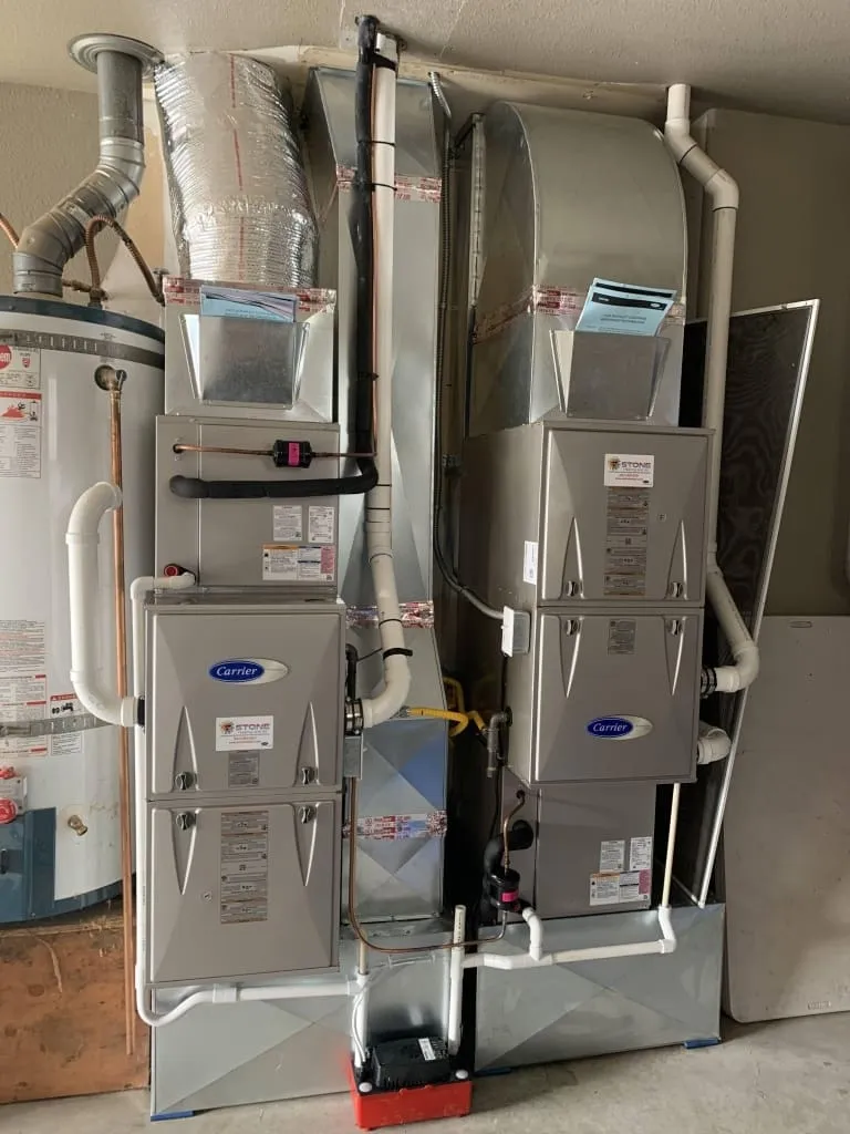Two gray Carrier air conditioning units with connected ductwork and piping installed side by side in a utility room.