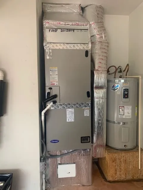 Vertical HVAC unit with a Carrier logo, insulated ductwork, and a water heater mounted on a wooden platform in a utility room.