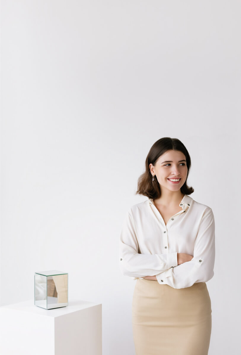 Young woman with short brown hair smiling and standing with arms crossed next to a mirrored cube on a white pedestal against a white background.