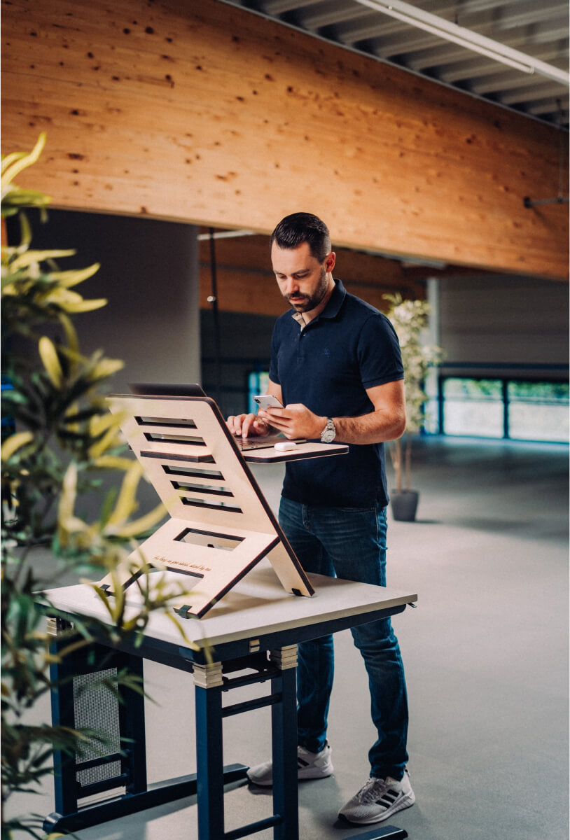 Man in navy blue polo shirt standing at a wooden standing desk using a laptop and holding a smartphone in a spacious office.