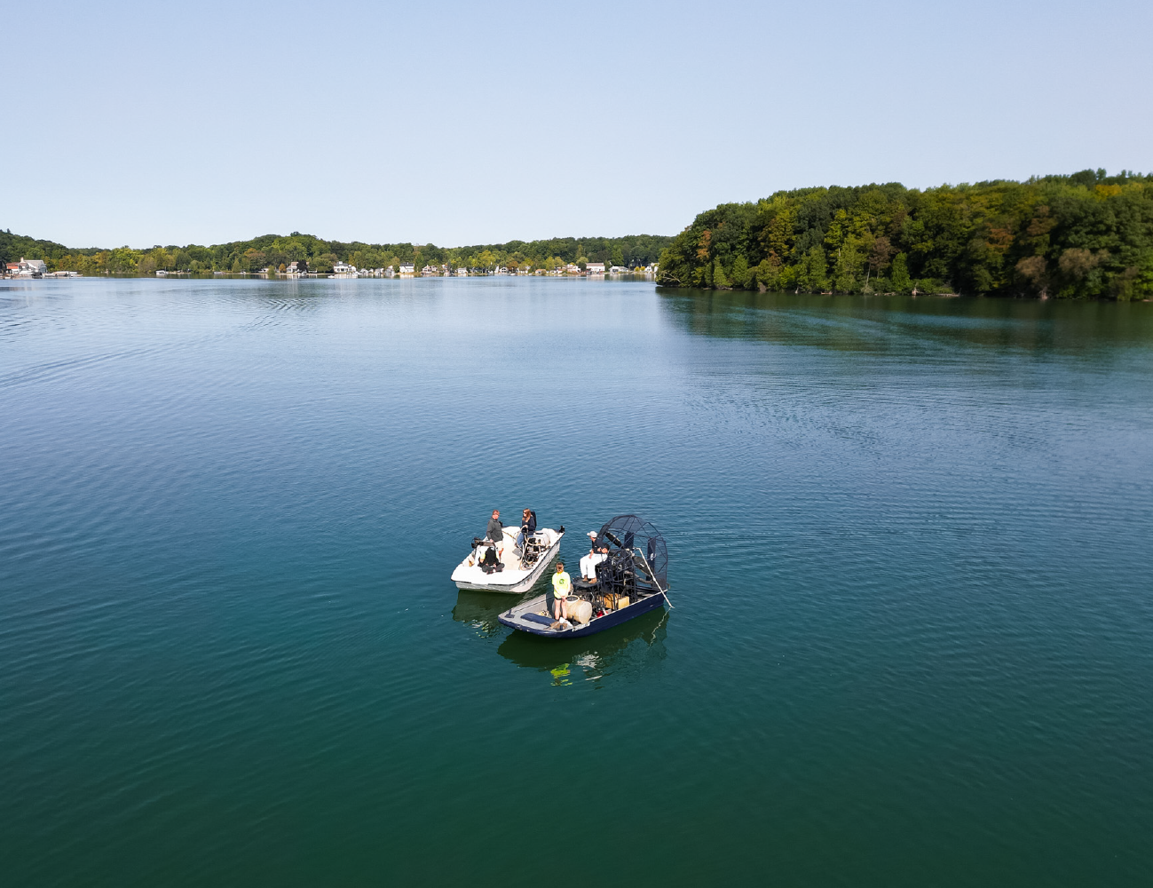 Two boats with people floating on a calm lake near a forested shoreline and distant houses.