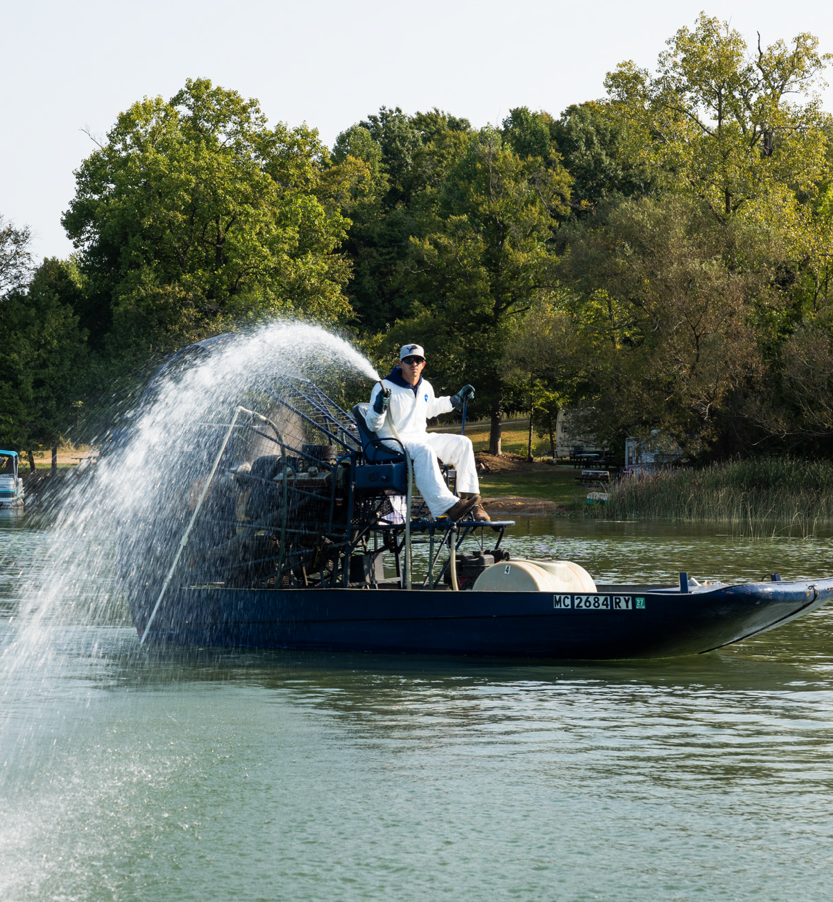 Man in white protective suit spraying water from an airboat on a lake near a tree-lined shore.