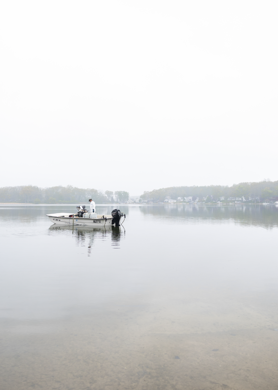 Person standing on a small boat in calm water near a shoreline with houses and trees under a cloudy sky.