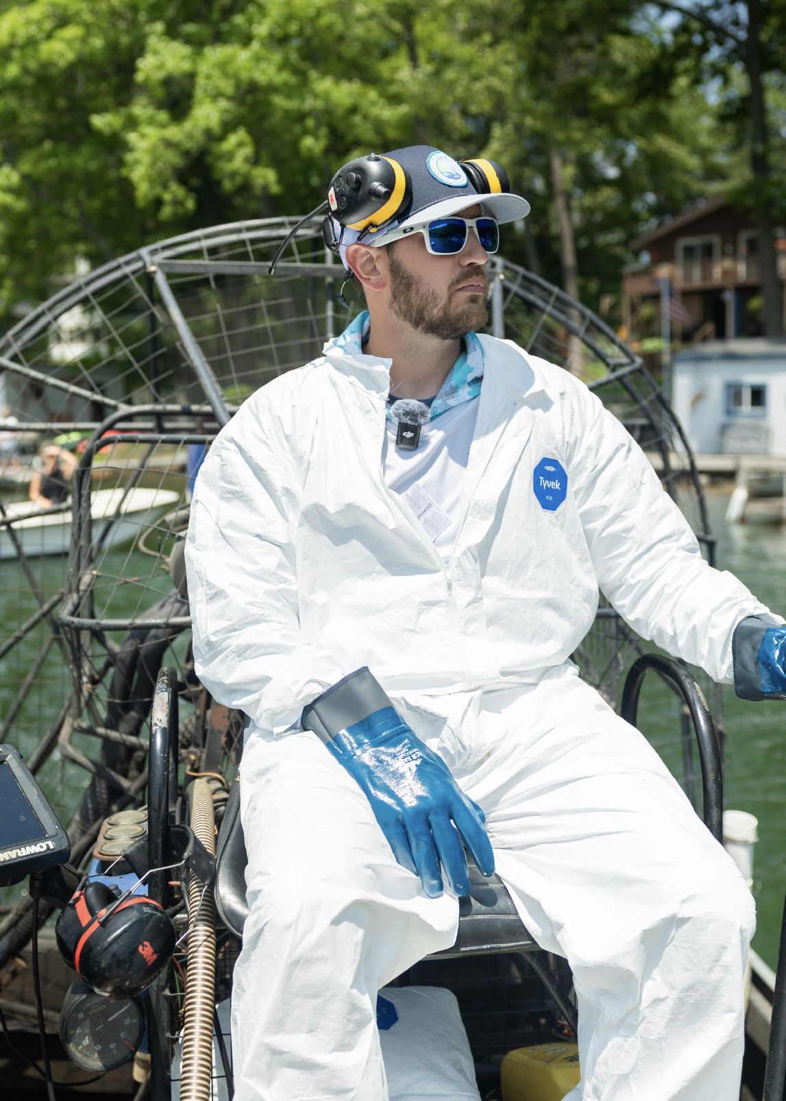 Man wearing white protective Tyvek suit, blue gloves, and a hat with earmuffs, sitting on an airboat near water with trees and houses in the background.