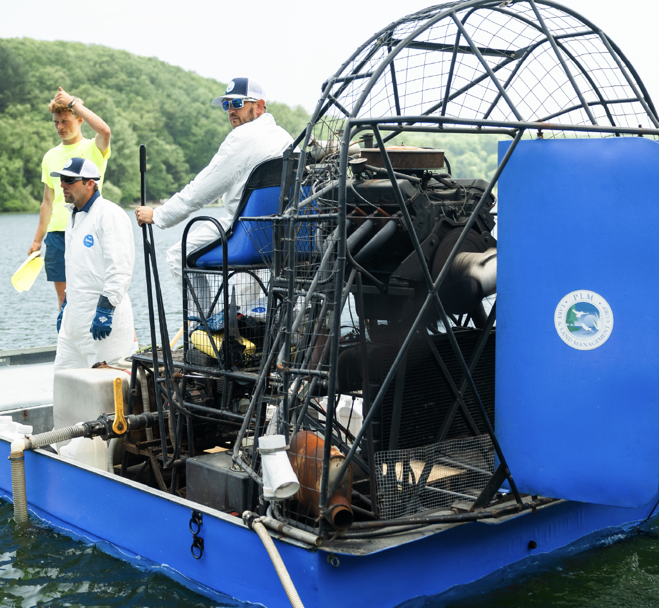 Three men on a blue airboat on a lake surrounded by forest, two men in white protective suits and one holding a yellow paddle.