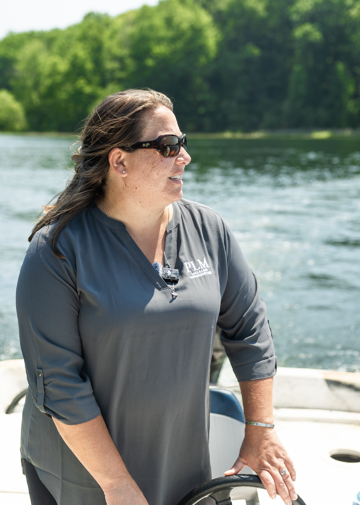 Woman wearing sunglasses and a PLM shirt steering a boat on a lake with green trees in the background.