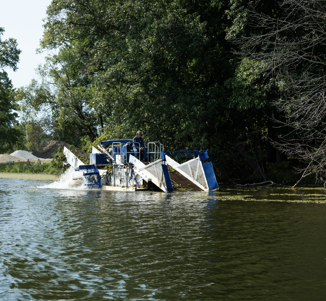 Man operating a blue and white aquatic weed harvester on a lake surrounded by trees.
