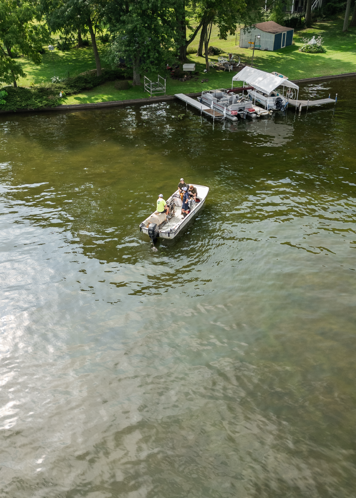 A small motorboat with five people on board floating on green lake water near a dock and a grassy shore with trees and a small shed.