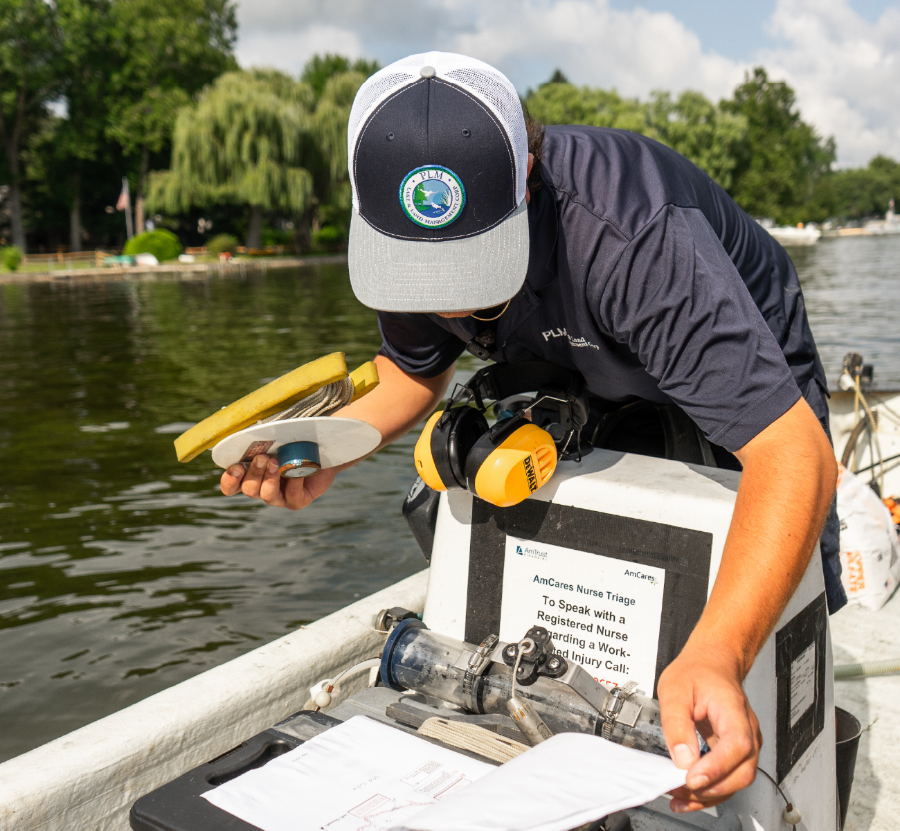 Person wearing a PLM cap and shirt working from a boat on a lake, handling water sampling equipment and documents.