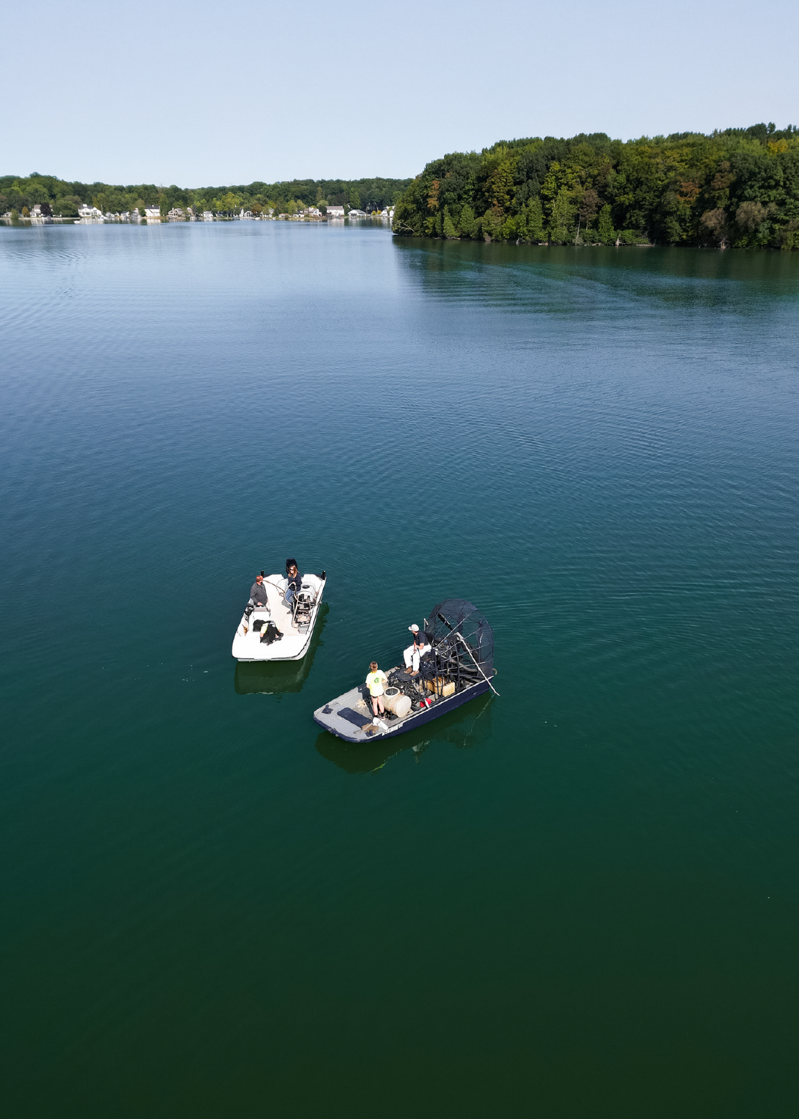 Two boats on calm water near a forested shoreline with houses in the distance under a clear sky.