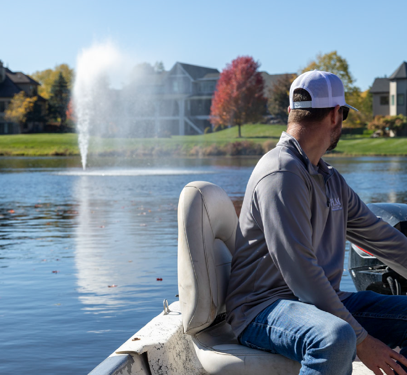Man wearing a white cap and sunglasses sitting in a boat on a lake with a water fountain in the background and houses along the shore.