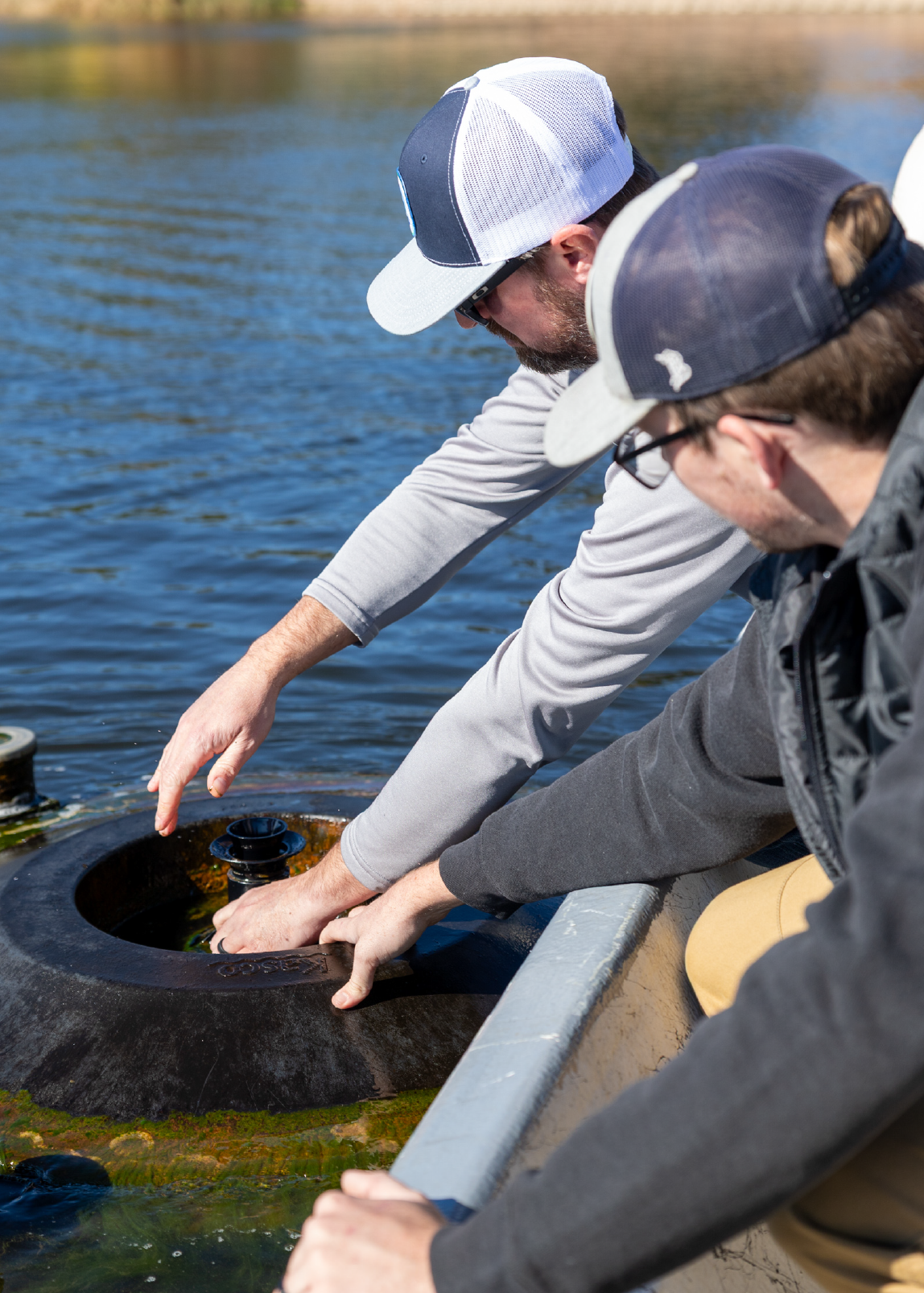 Two men wearing caps adjusting a fountain device partially submerged in a pond from the edge of a boat.