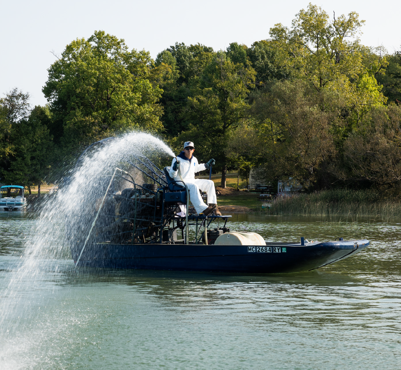 Person in white protective suit operating airboat spraying water over a lake near a tree-lined shore.