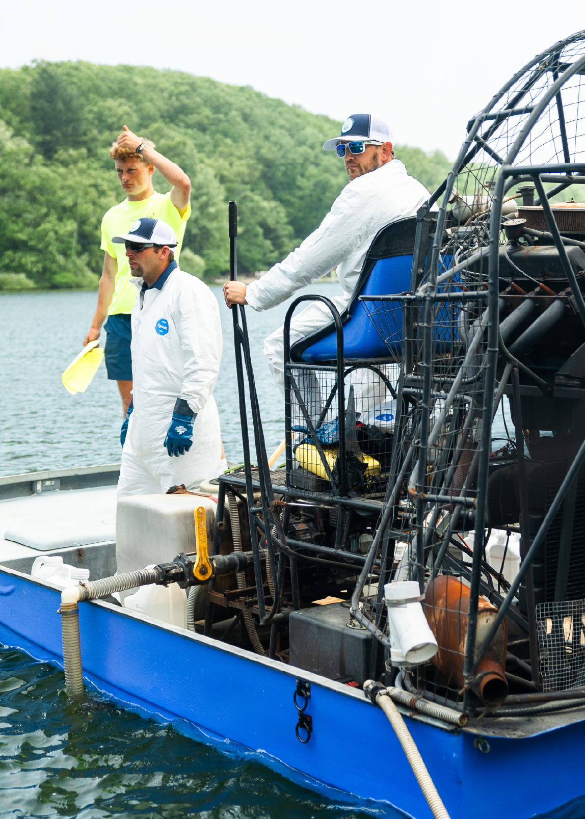 Three men on a blue airboat on a lake, with two men wearing white protective suits and caps, and one man in a yellow shirt holding yellow paddles.