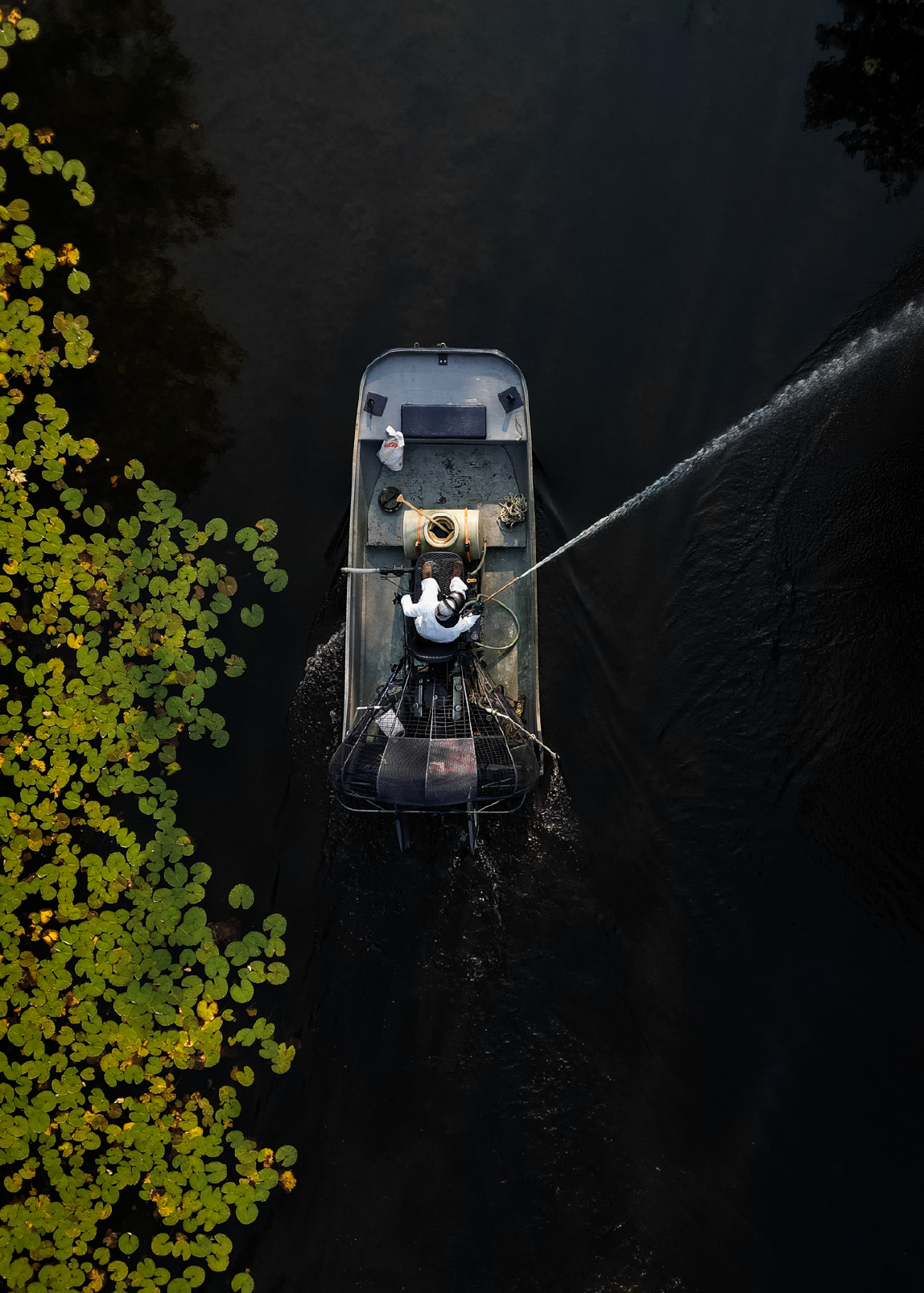A person in protective clothing operates a machine on a boat spraying water over dark river water next to floating green lily pads.