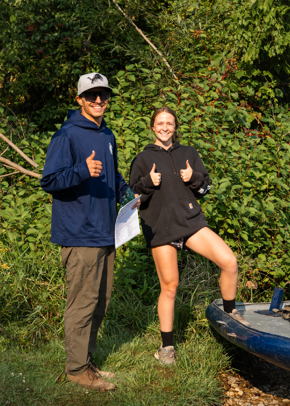 Two people outdoors smiling and giving thumbs up, one standing on grass holding a paper and the other with one foot on a blue boat.