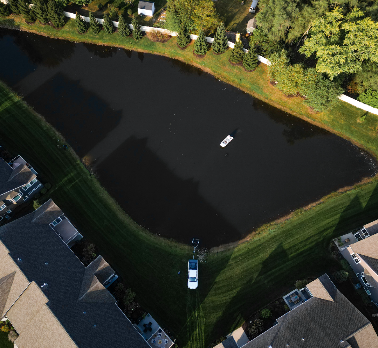 Aerial view of a small rectangular pond surrounded by green lawns and residential houses, with a white vehicle parked near the pond's edge and a floating aerator in the water.