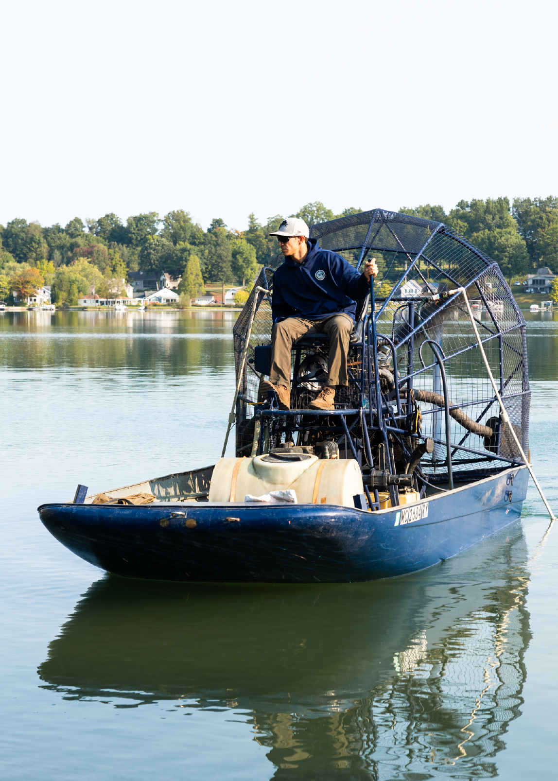 Man wearing sunglasses and cap operating a blue airboat on calm water with houses and trees in the background.