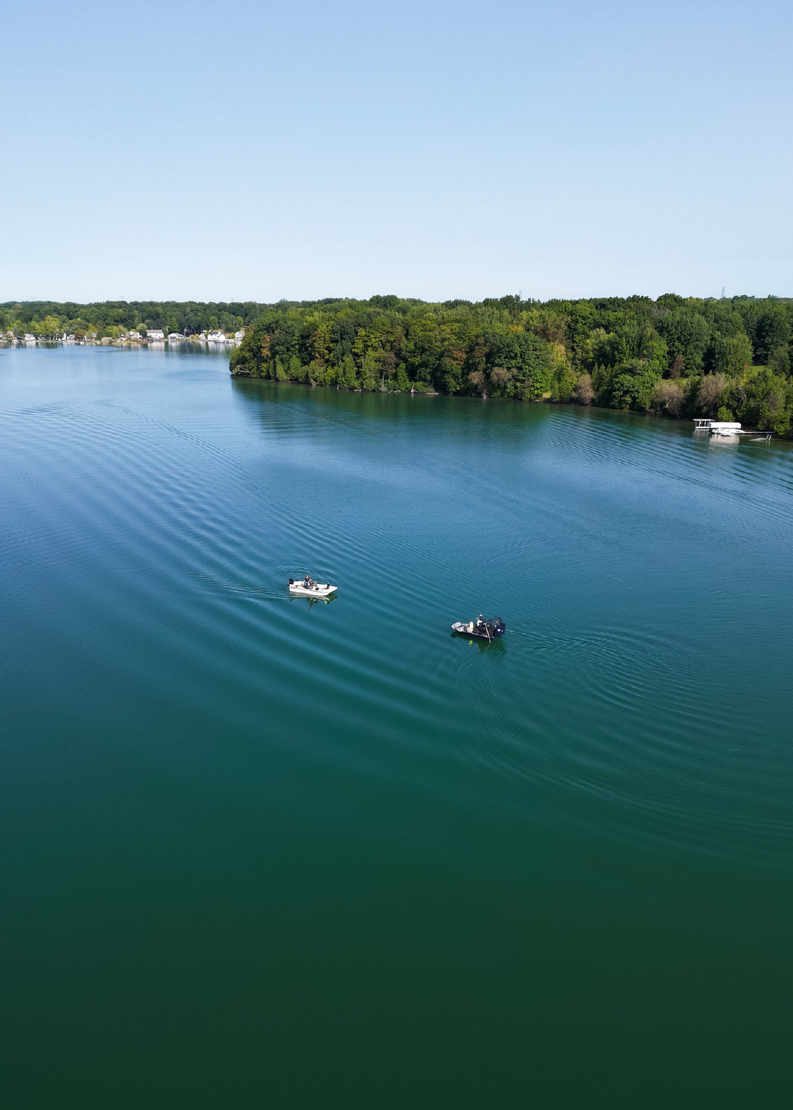 Two small boats on a calm lake with green forested shoreline under a clear blue sky.