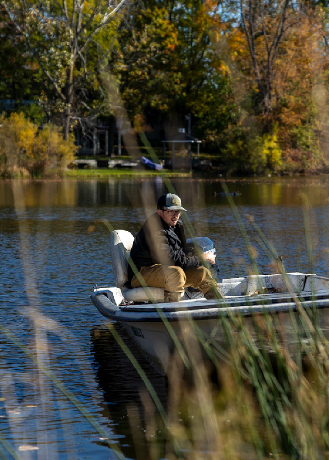 Man wearing a cap and jacket sitting in a small motorboat on a calm lake with autumn trees in the background.