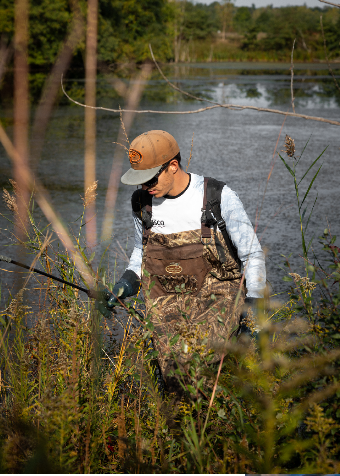 Man wearing camouflage waders, a white shirt, and a brown cap standing in tall grass near a body of water holding a tool.