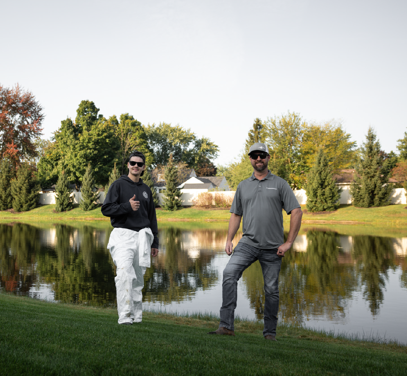 Two men standing on green grass near a calm pond with trees and houses in the background.