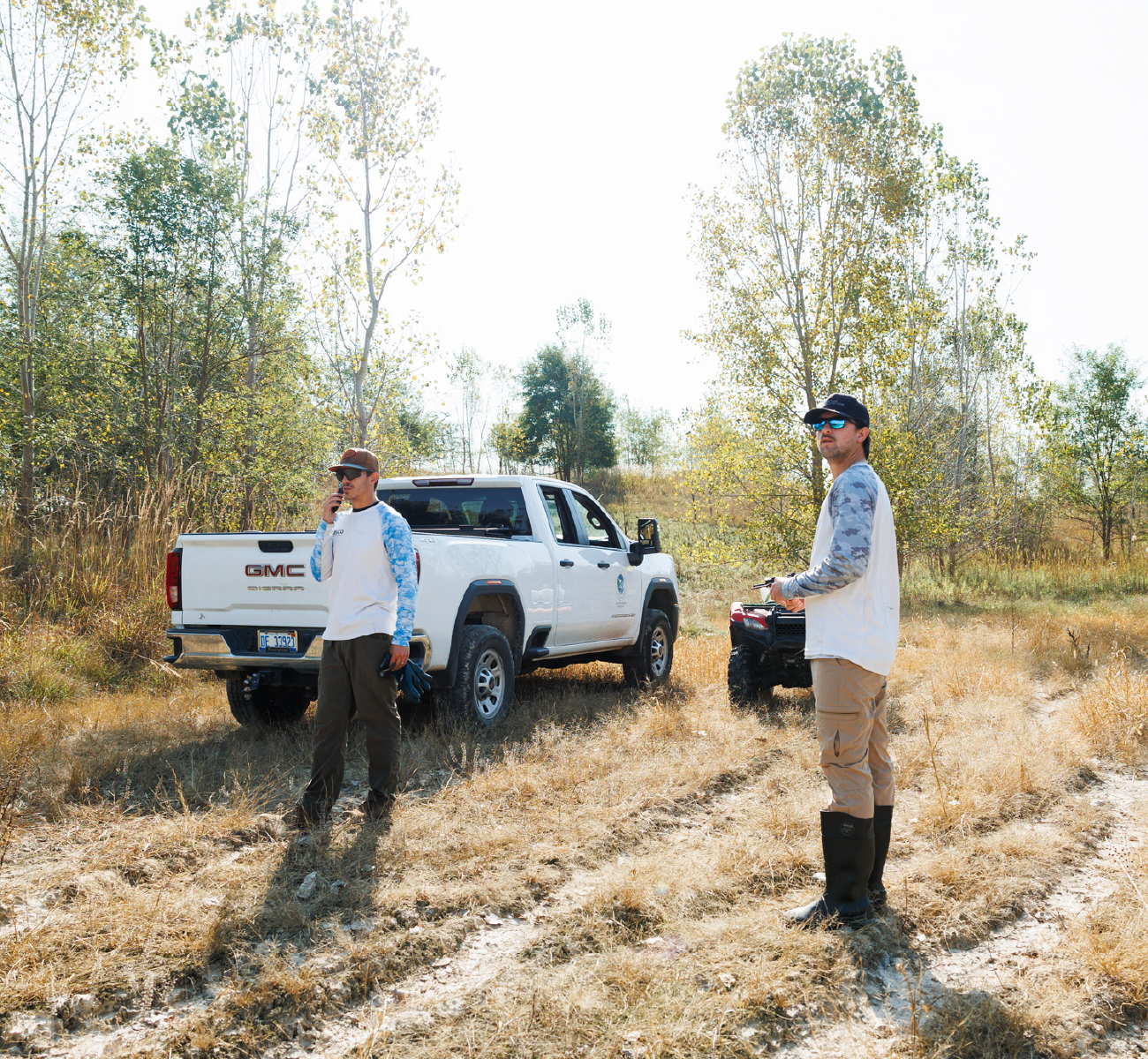 Two men wearing sunglasses and casual outdoor clothing standing near a white GMC pickup truck and an ATV in a grassy area with sparse trees.