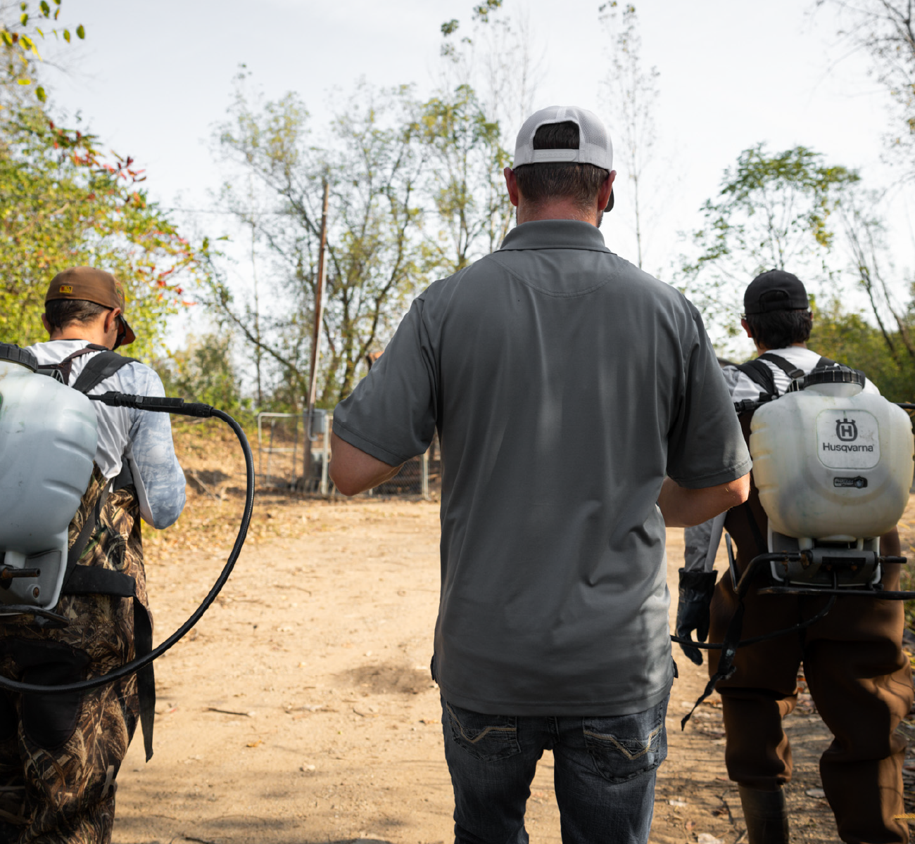 Two people wearing backpack sprayers walk beside a man in a gray shirt and white cap outdoors on a dirt path with trees in the background.