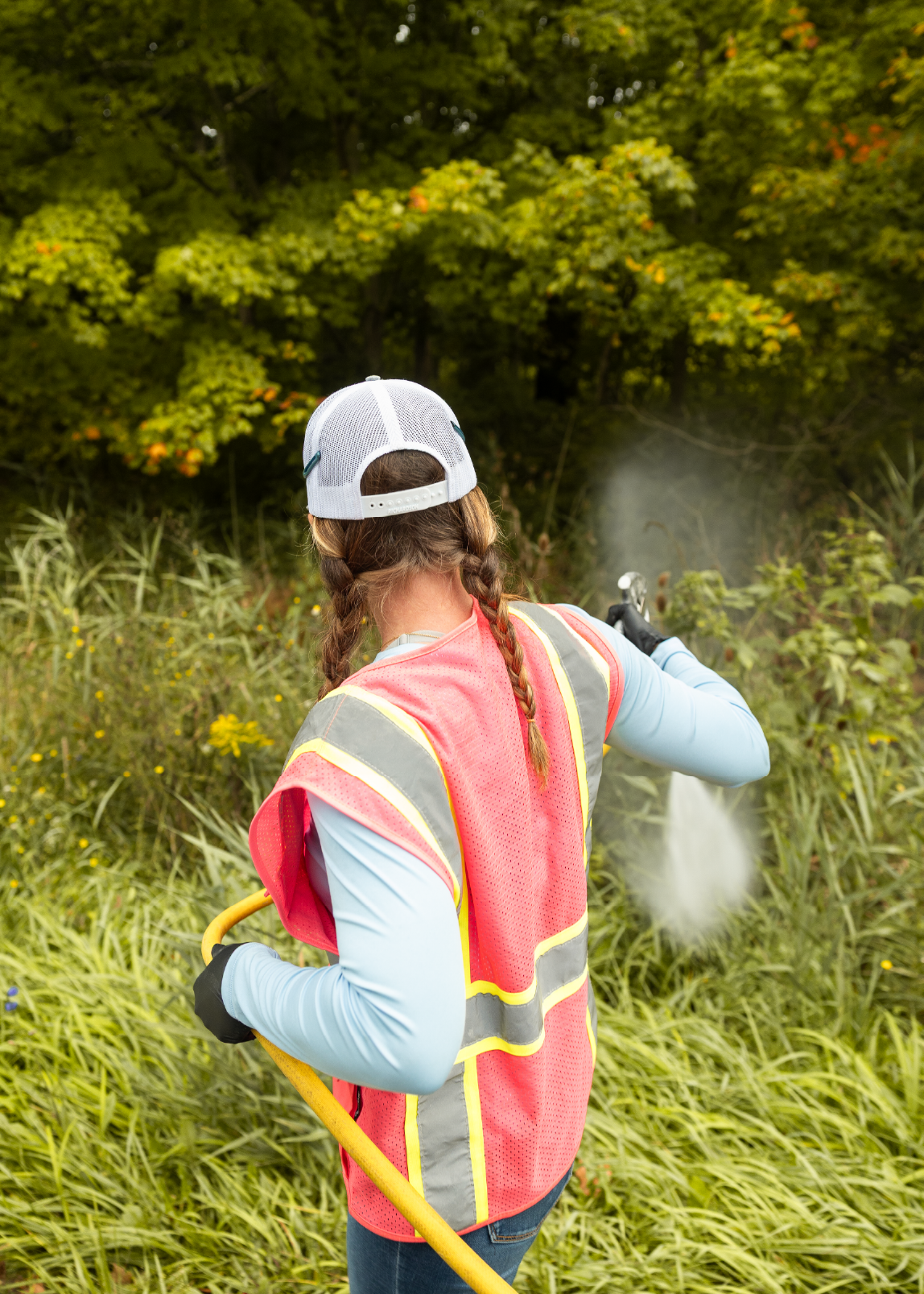 Person in a white cap and orange reflective vest spraying herbicide on plants with a hose in a green outdoor area.