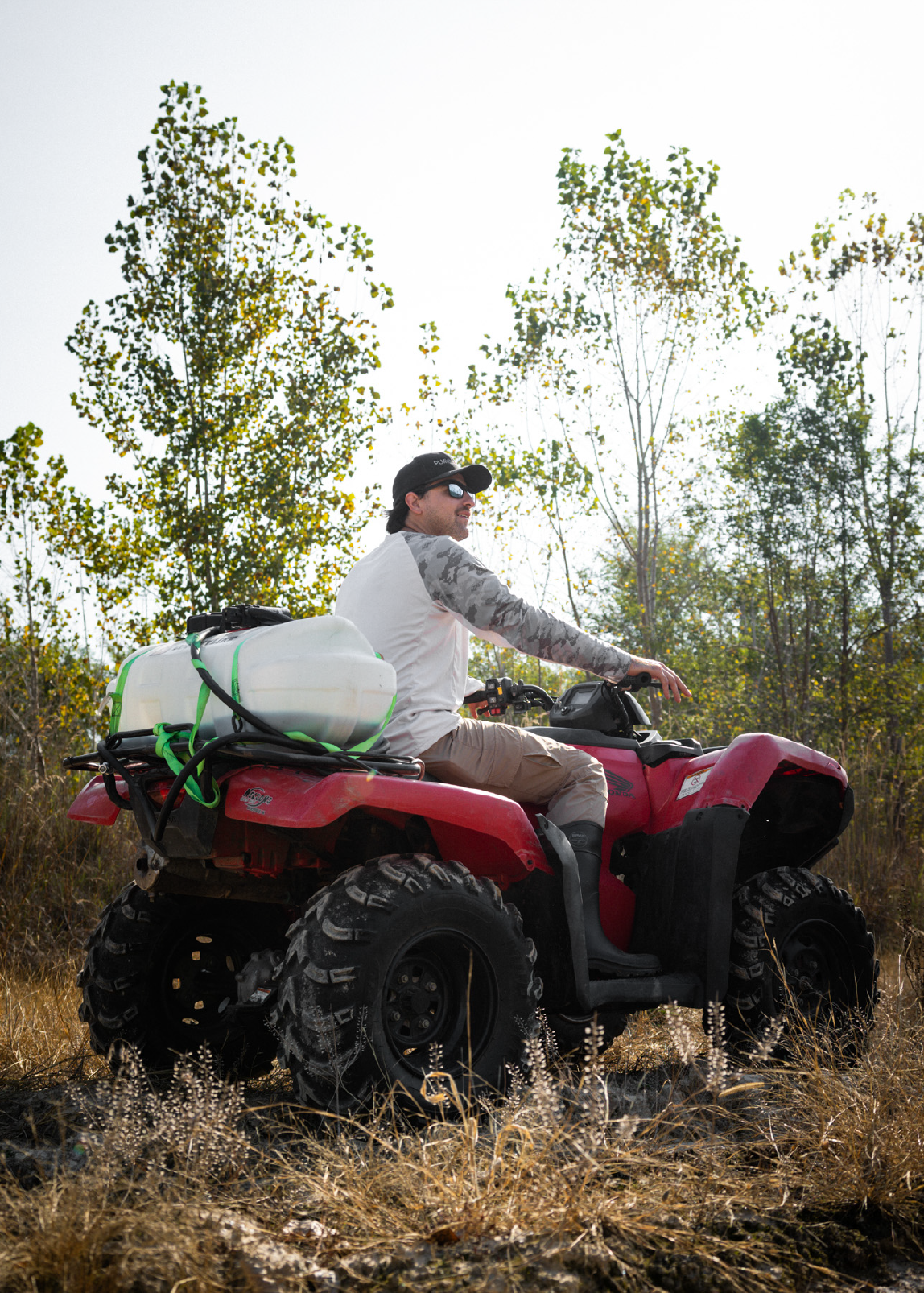 Man wearing sunglasses and cap riding a red ATV in a grassy area with a large white tank strapped to the back.