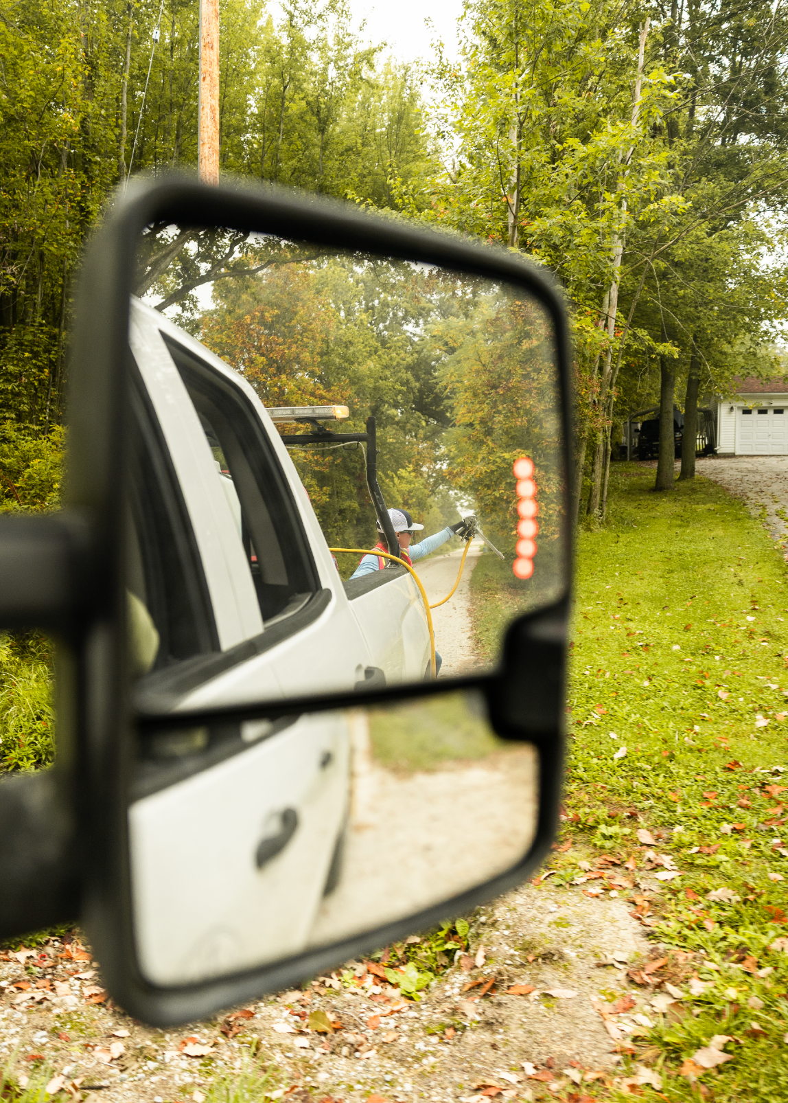 Side mirror view of a worker spraying vegetation along a rural roadside from a white pickup truck.