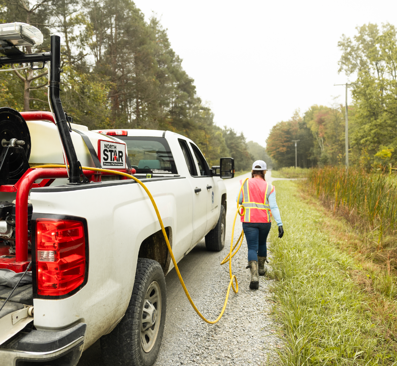 Worker in safety vest walking along roadside gravel path next to a white truck with vegetation control equipment.