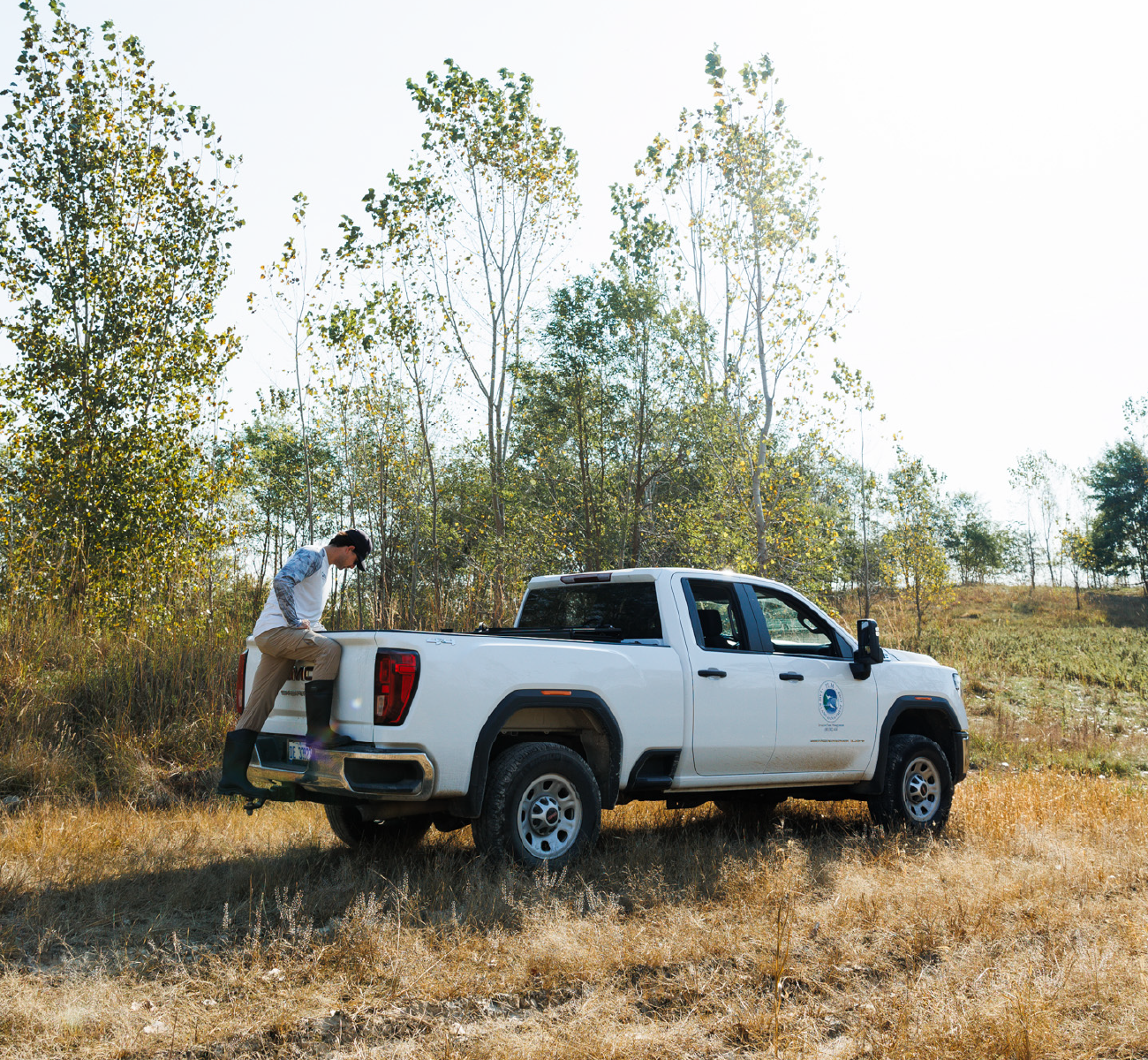 Person in tan pants and rubber boots stepping into the bed of a white pickup truck parked in a field with sparse trees in the background.