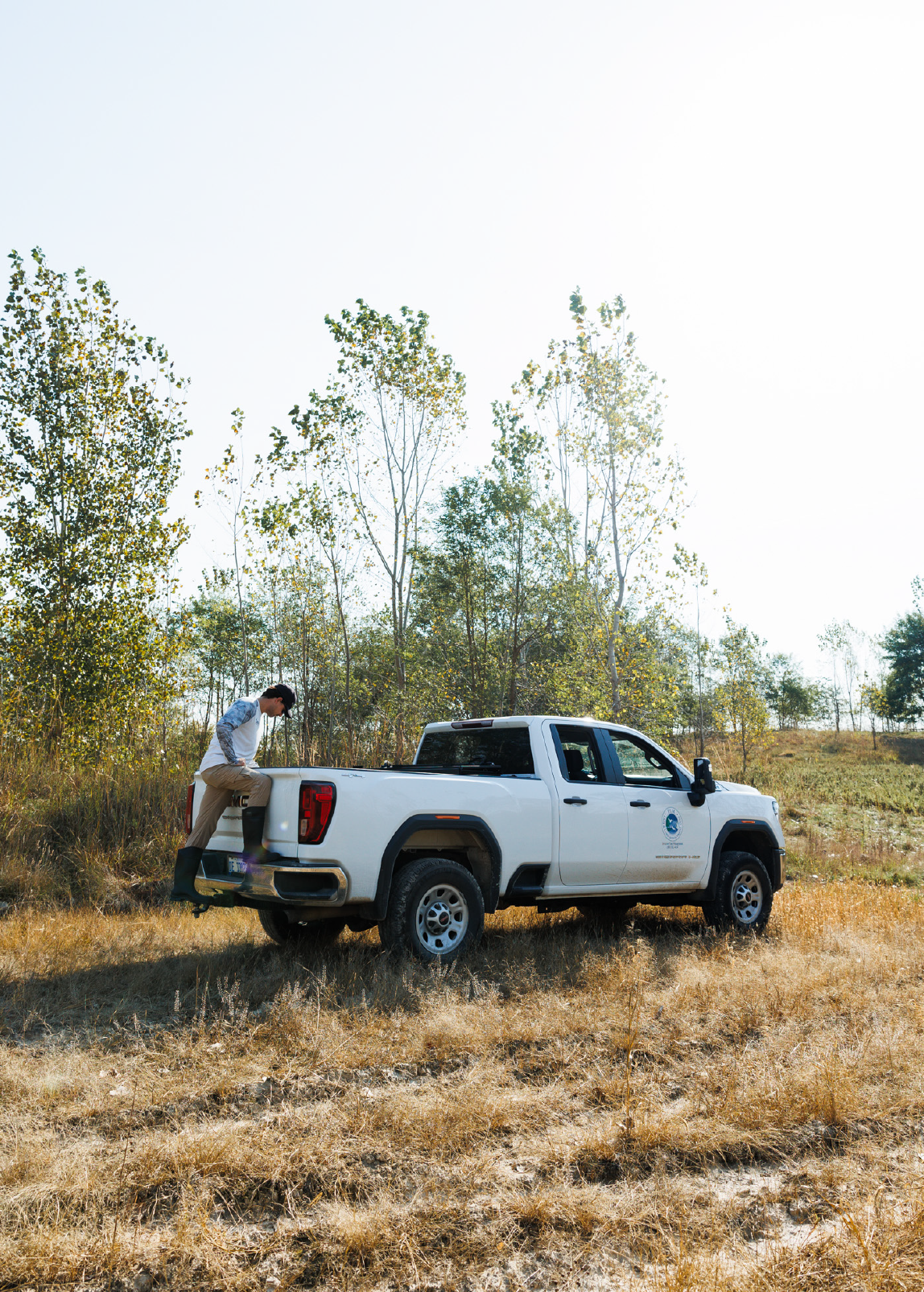 Person wearing rubber boots stepping into the bed of a white pickup truck parked on dry grass near tall trees.