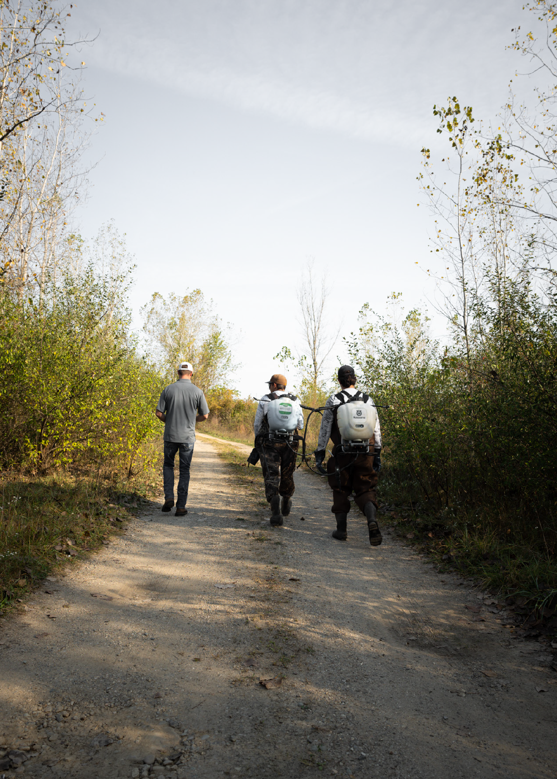 Three people walking on a dirt path through a wooded area, two wearing backpack sprayers.