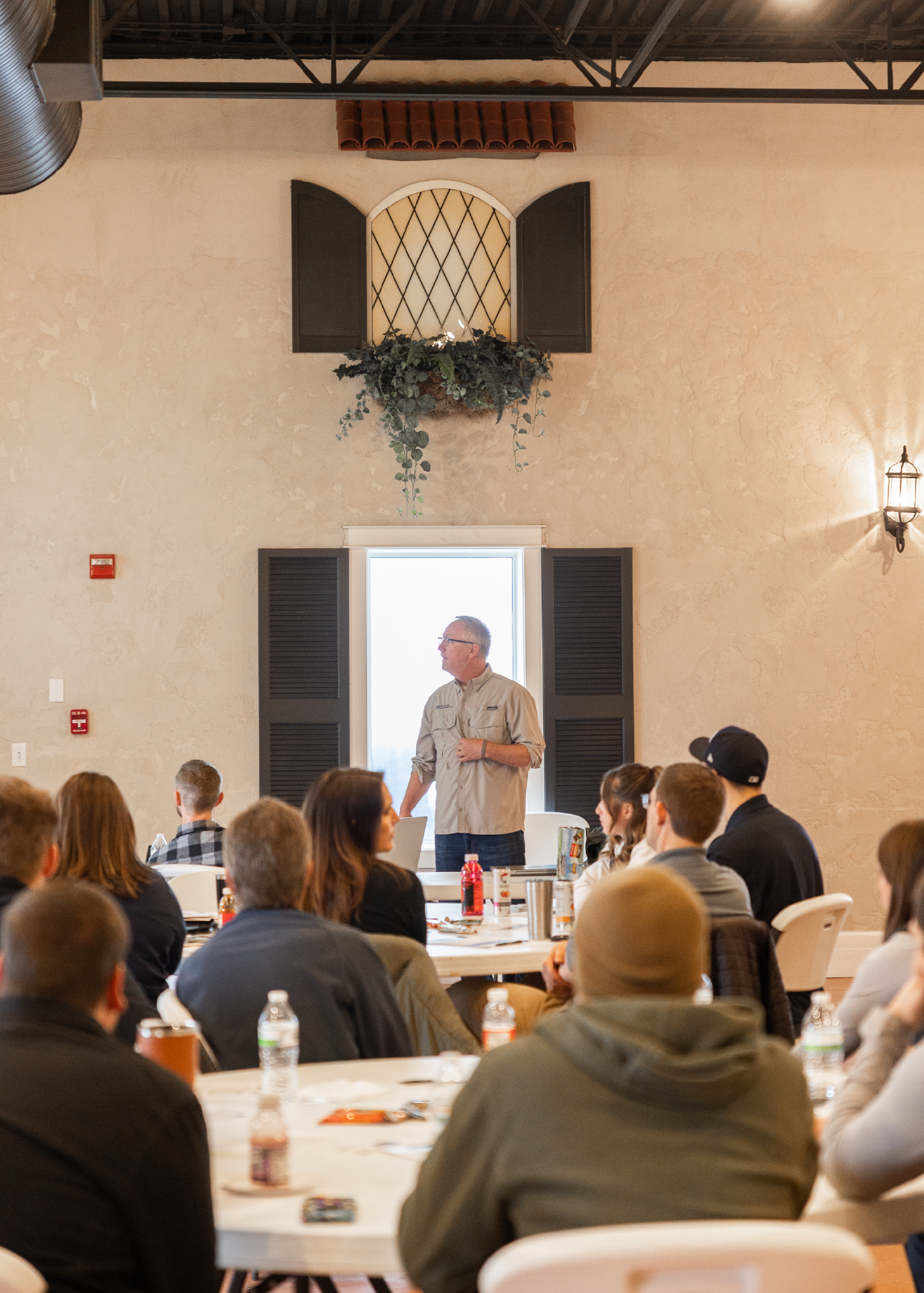 Man giving a presentation to an audience seated at round tables in a rustic room with a window and decorative plants.