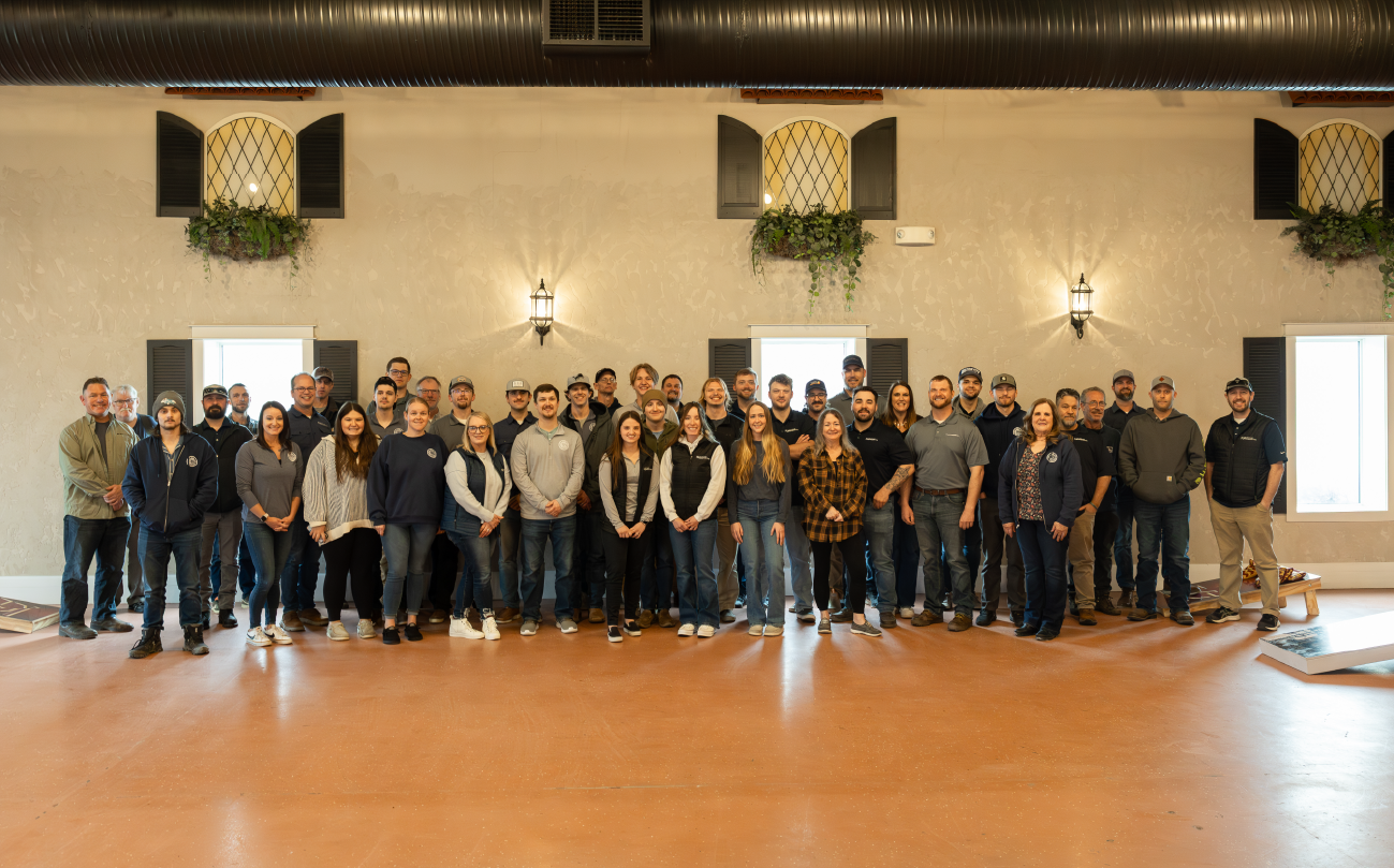 Large group of diverse men and women standing indoors in front of a beige wall with two windows and decorative plants.