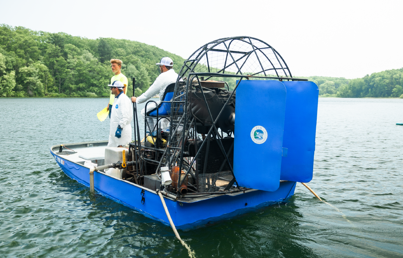 Three men on a blue airboat on a lake surrounded by green trees.