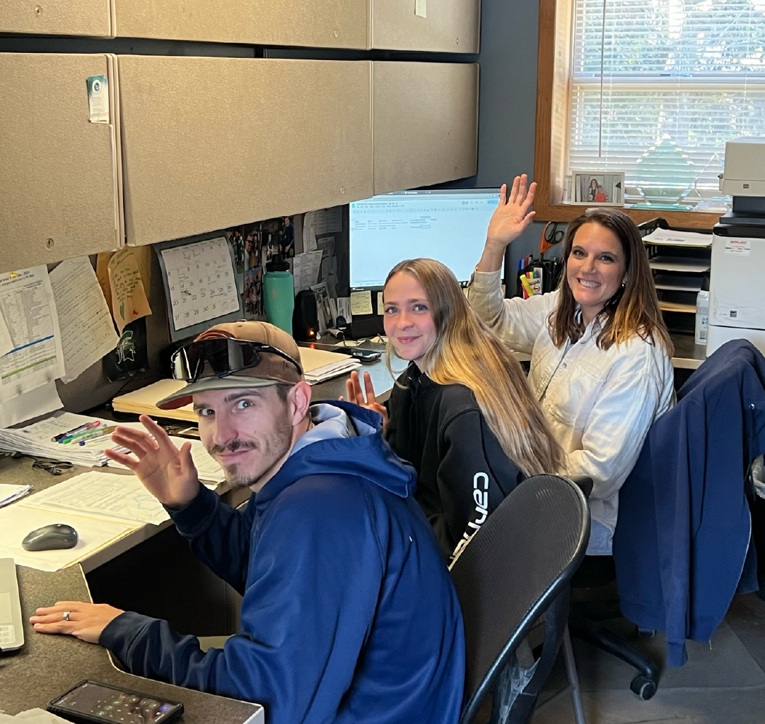 Three smiling people sitting closely at an office desk waving at the camera, with computers, papers, and office supplies around them.