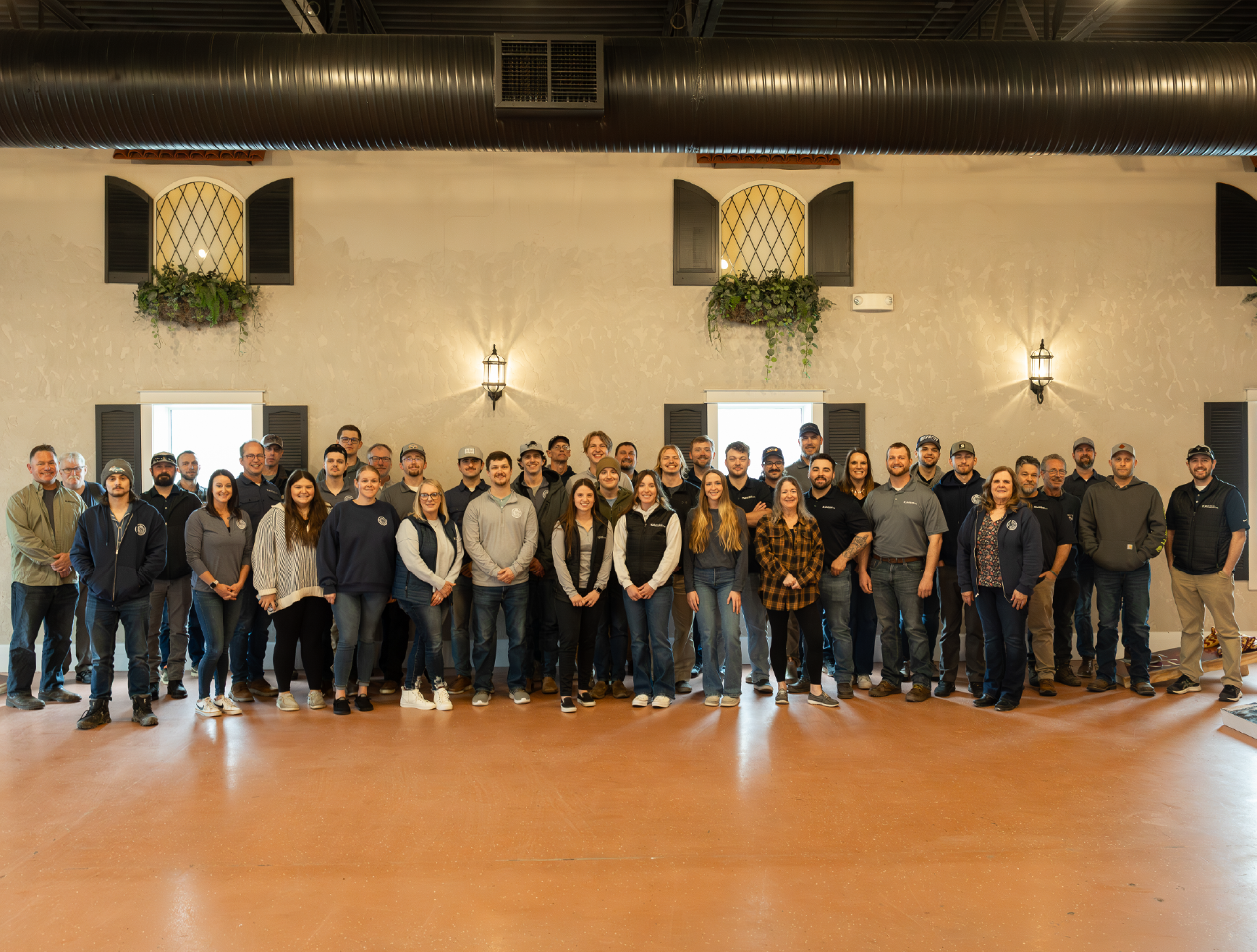 Large group of people standing indoors in two rows in front of a beige wall with two windows and wall sconces.