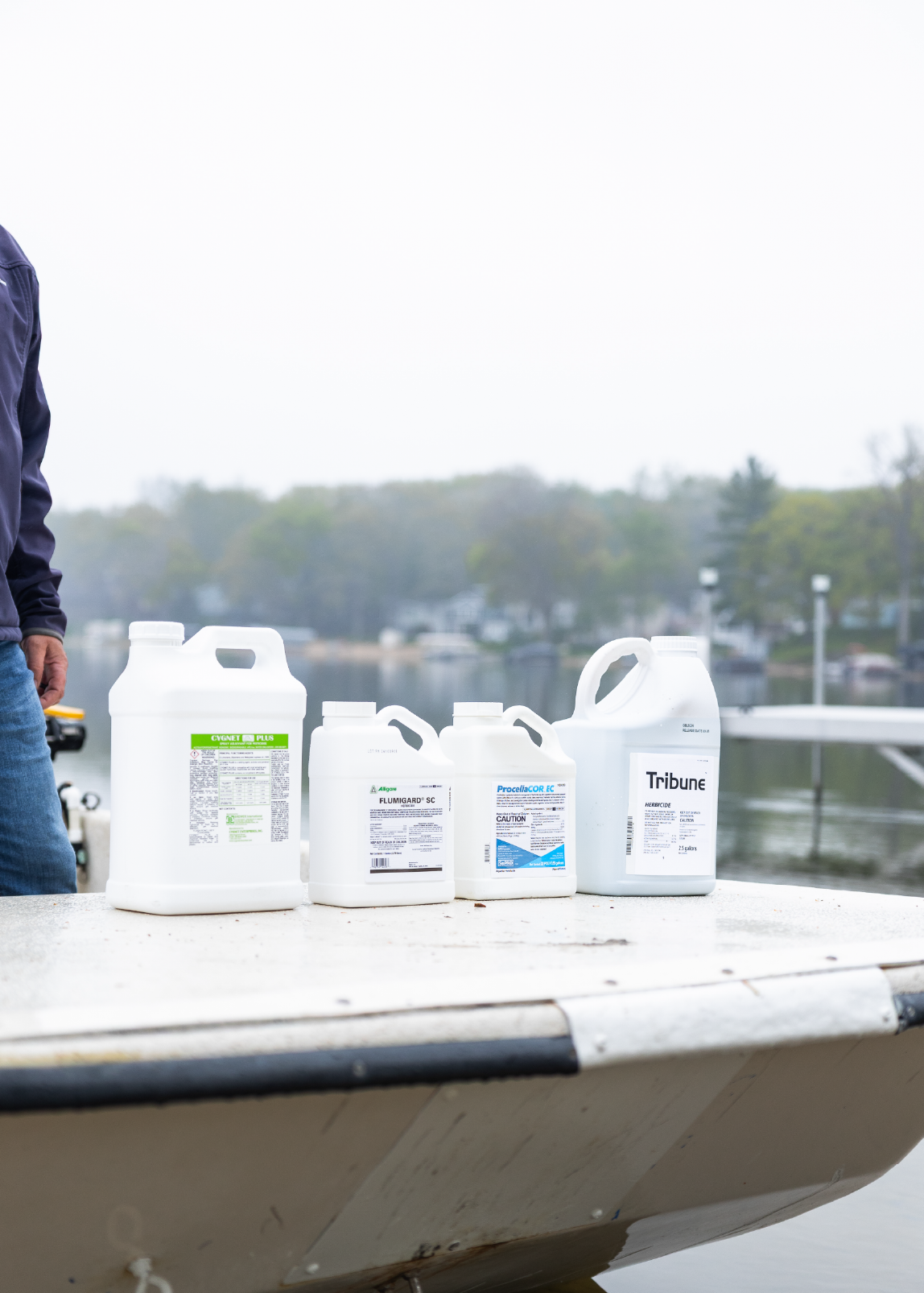 Four white chemical containers labeled Cygnet Plus, Flumigard SC, Procelia COR EC, and Tribune on a boat near a lakeside dock.