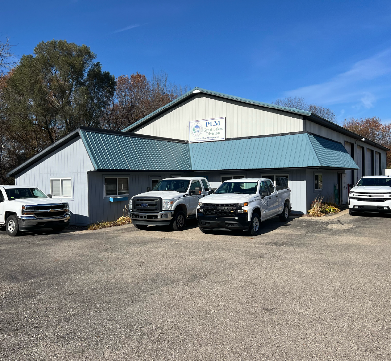 Office building with a blue metal roof and a sign reading PLM Great Lakes Division, with four parked pickup trucks in front.