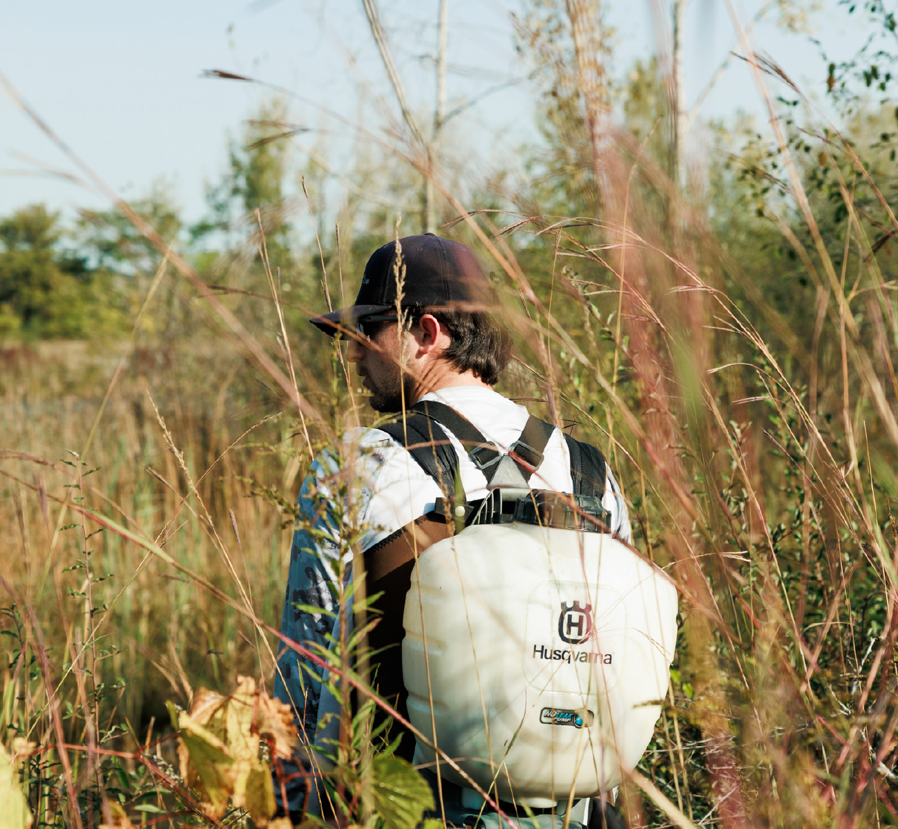 Person wearing a black cap and glasses with a white Husqvarna backpack sprayer in a tall grassy field.