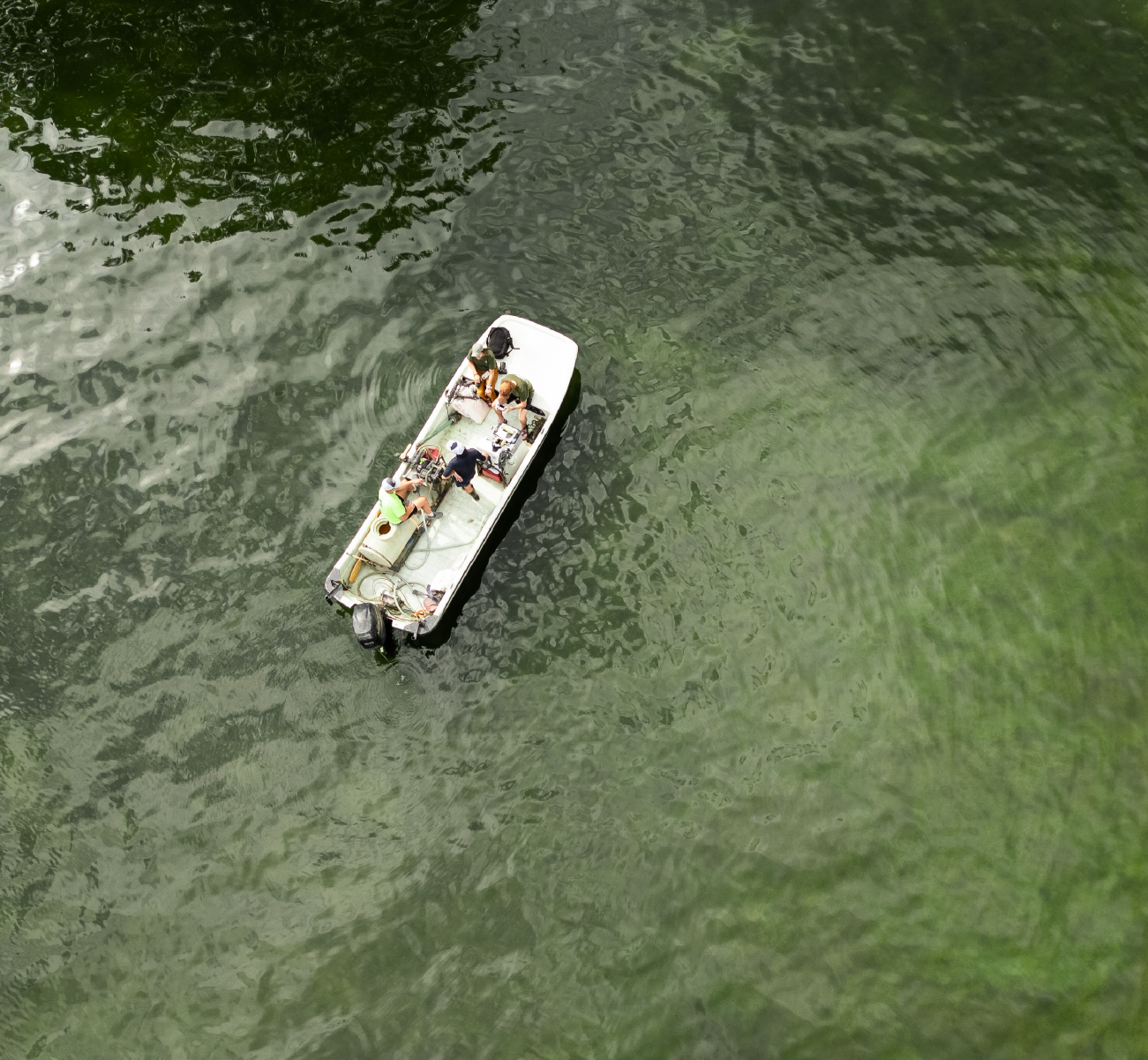 Aerial view of a small boat with four people working on equipment on calm green water.