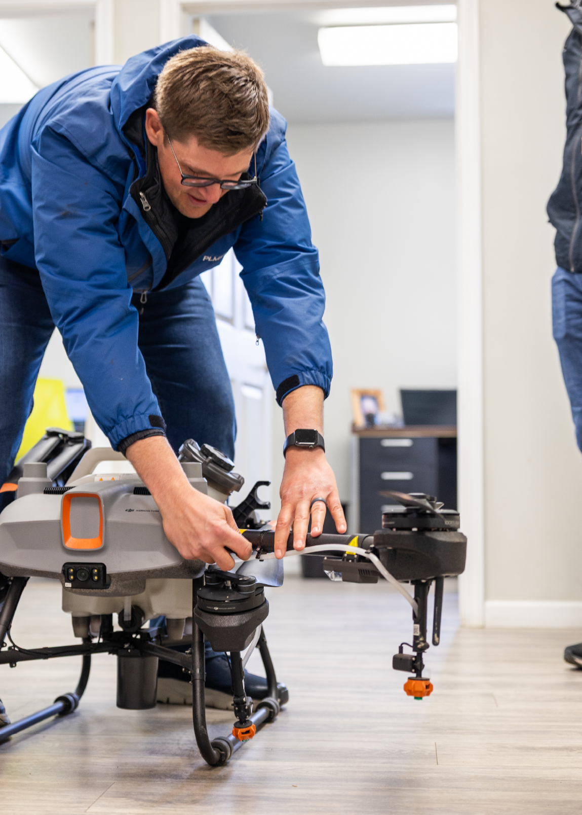 Man in a blue jacket adjusting components on a large professional drone indoors.